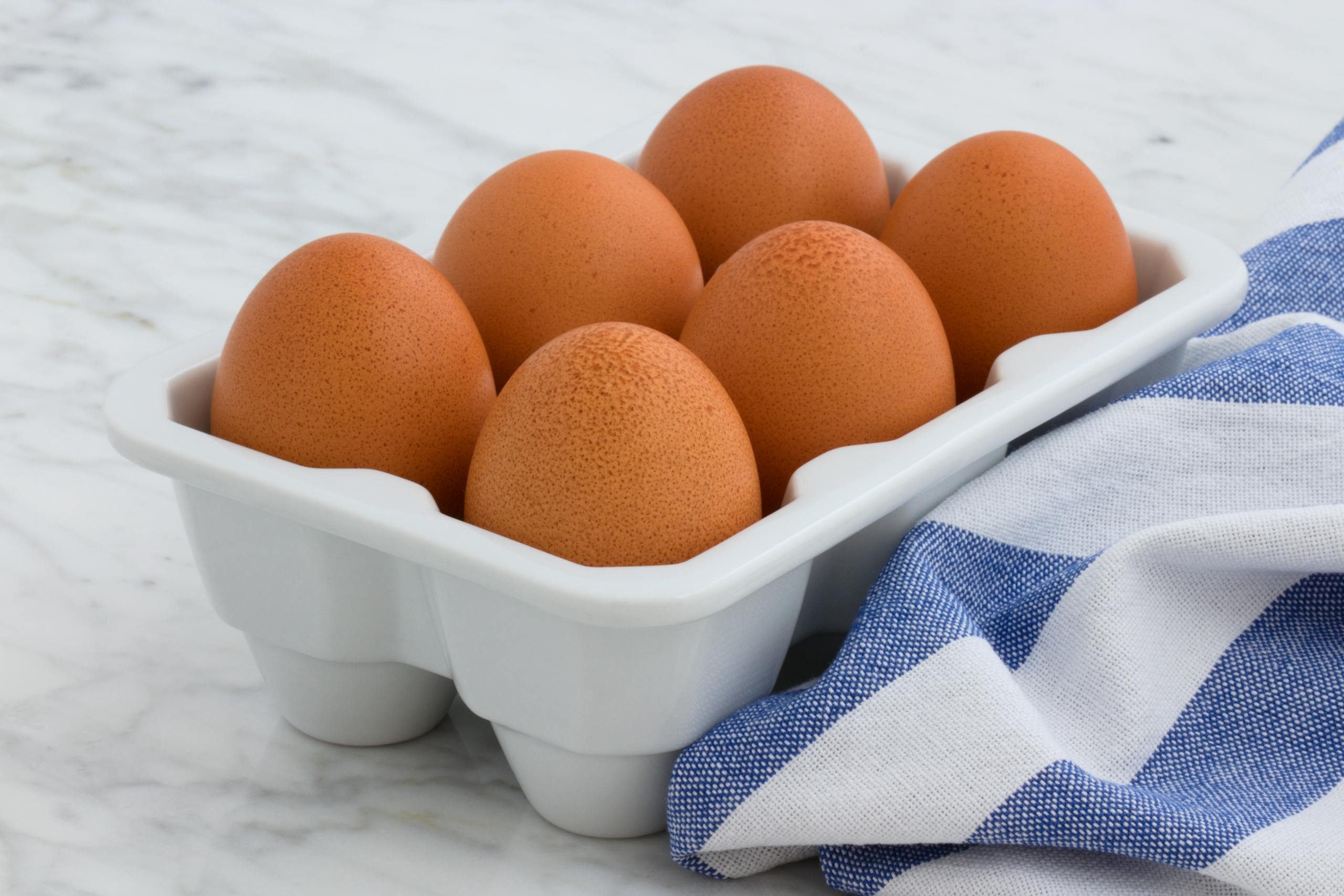 A close-up of fresh brown eggs arranged in a ceramic tray with a striped cloth on a marble surface.