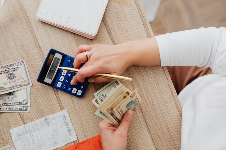 Person using a calculator and counting cash on a wooden table with receipts.