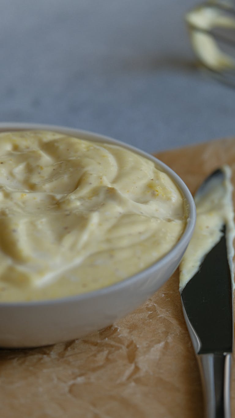 A bowl of creamy homemade mayonnaise on a cutting board with a spreading knife.