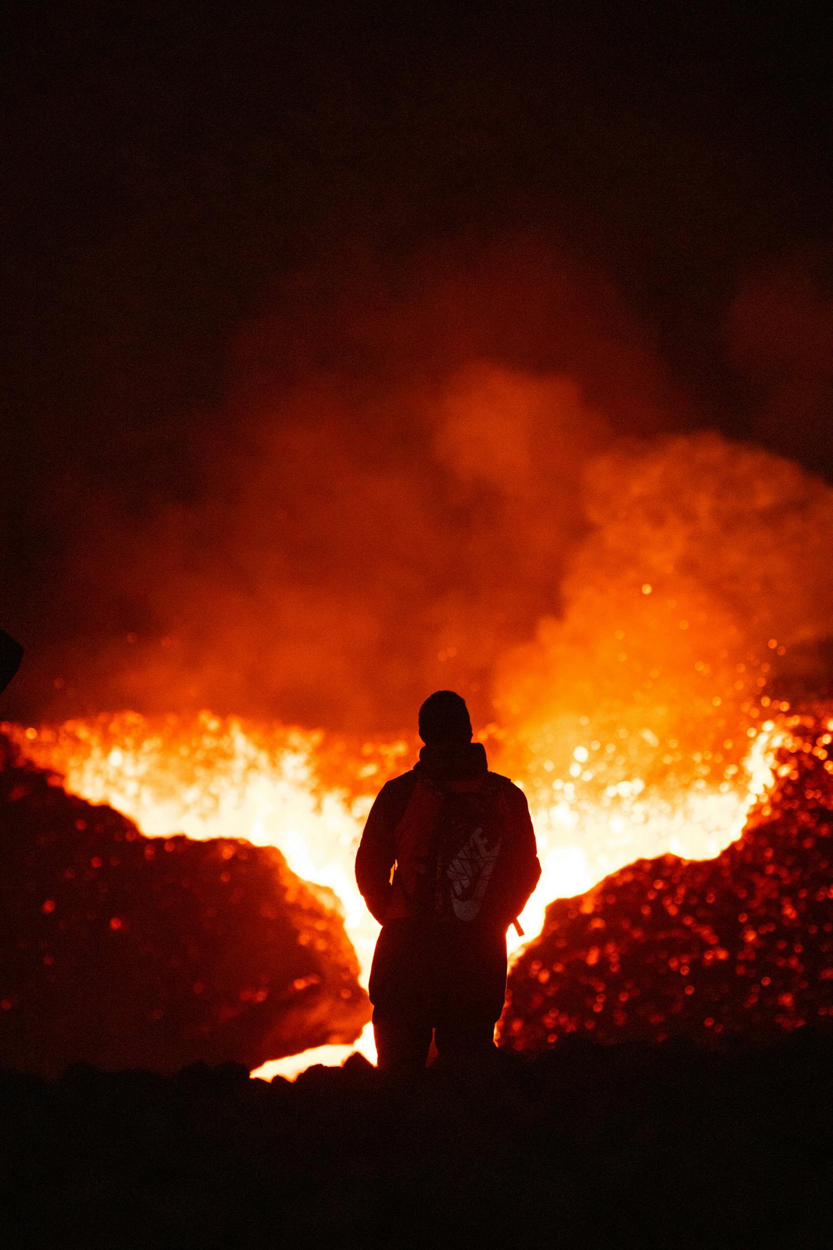 A person silhouetted against a fiery volcanic eruption, observing the intense lava flow.