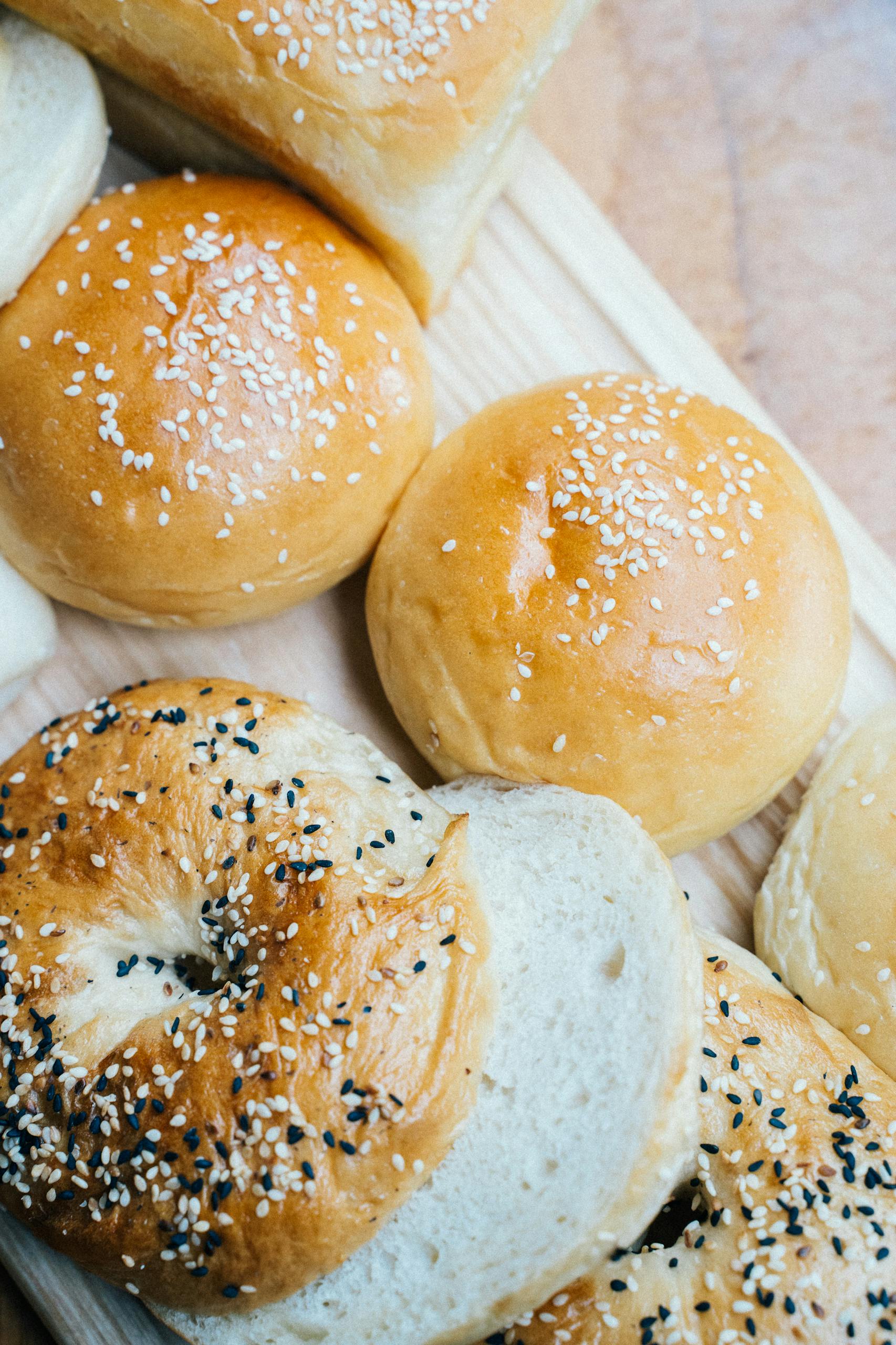 Close-up of freshly baked bagels and buns topped with sesame seeds on a wooden board.